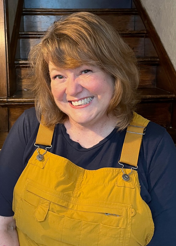 Picture of Trish Berrong, a smiling woman wearing a blue t-shirt and yellow overalls, sitting on the stairs because that's where the light in her house is best.