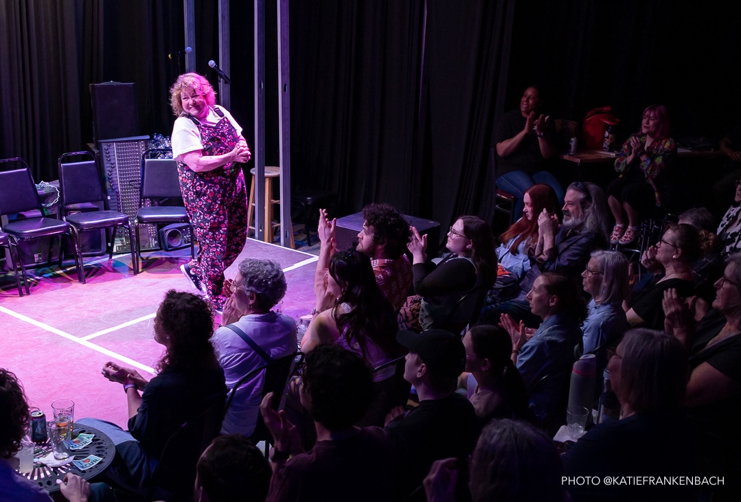 A woman in floral overalls stands on a stage in front of an applauding crowd.